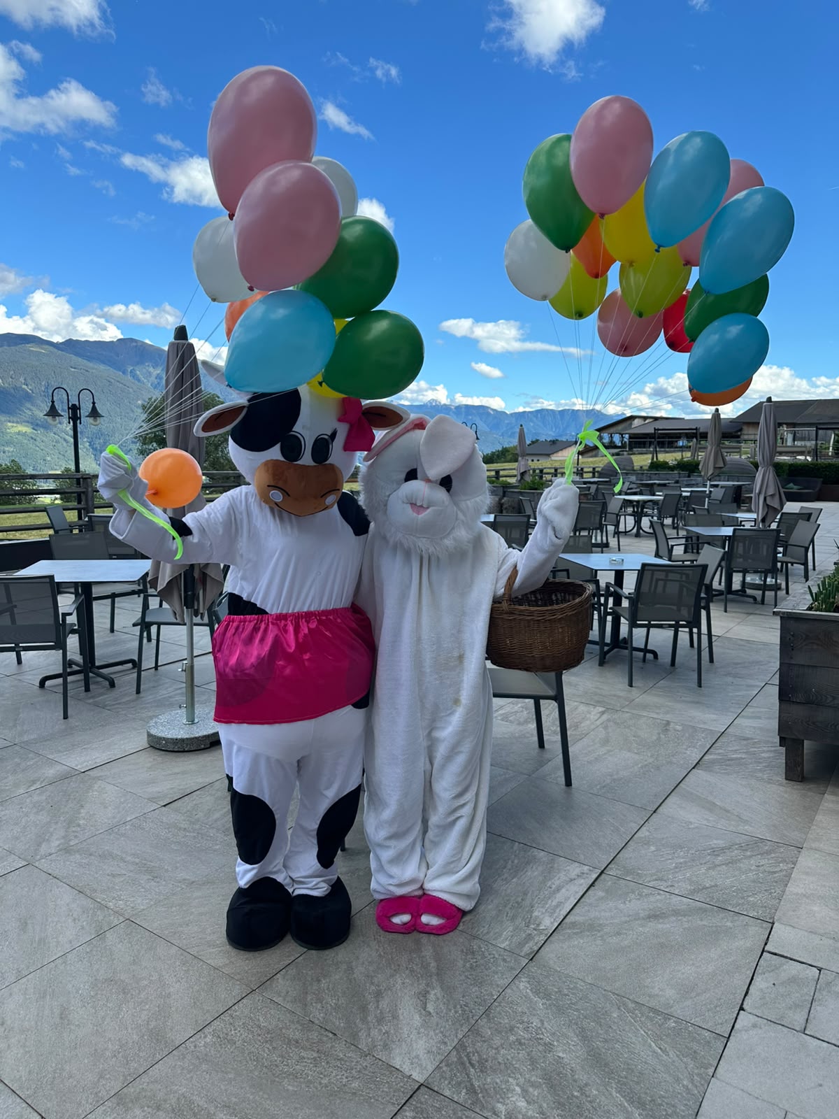 Zwei Hotelmaskottchen halten bunte Luftballons auf einer sonnigen Terrasse